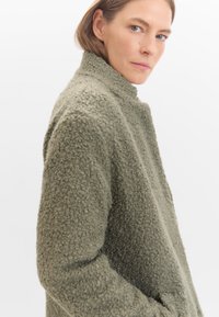 Olivgrüne, strukturierte Außenjacke mit hohem Kragen und lässiger Passform. Der Stoff hat eine weiche, flauschige Oberfläche.