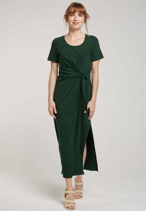 Robe verte foncée, jusqu'aux genoux, avec des manches courtes, présentant un nœud torsadé à l'avant et une fente sur le côté. Fabriquée en tissu lisse.