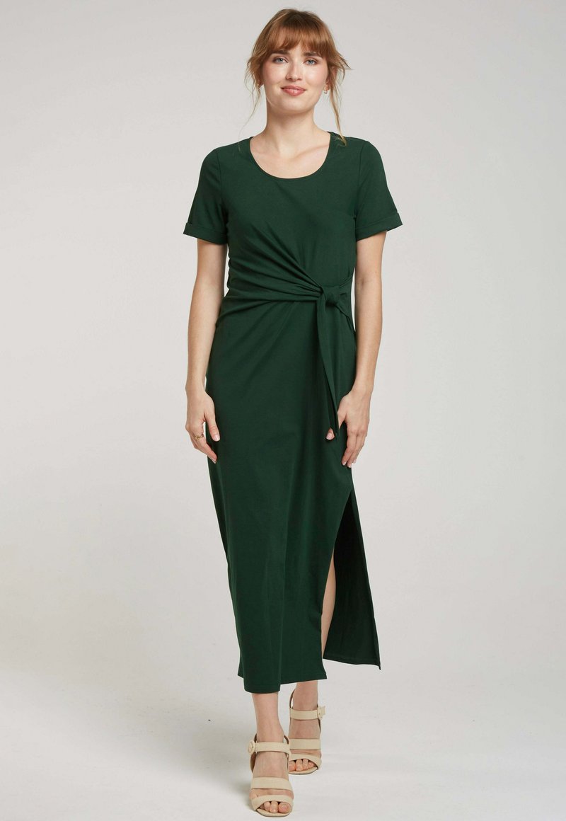Robe verte foncée, jusqu'aux genoux, avec des manches courtes, présentant un nœud torsadé à l'avant et une fente sur le côté. Fabriquée en tissu lisse.