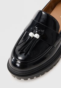 Mustat lakatunnahkakengät esittelevät rosoisen pohjan, leveän hihnan, kaksi helmeä ja tupsuja lisäyksinä. Sujuva ja kiiltävä pinta.