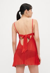 Punainen halkioinen mekko, jossa ohuet hihnat, avoin selkä ja suuri rusetti. Läpinäkyvää kangasta ja laskeutuva siluetti, joka antaa kevyen vaikutelman.