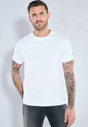 Weißes Baumwoll-T-Shirt mit Rundhalsausschnitt, kurzen Ärmeln und glatter Textur. Modell trägt schwarze Jeans und zeigt detaillierte Tattoos an den Armen.