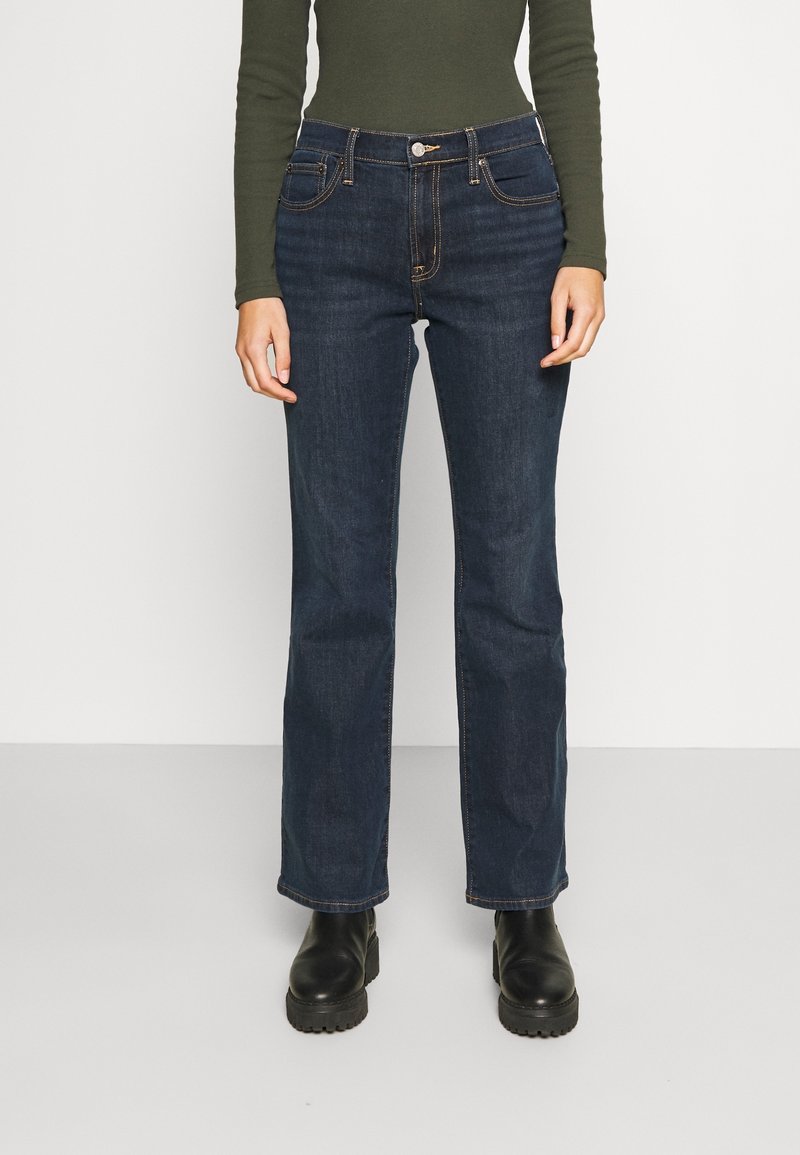 Personne portant un jean droit bleu foncé, un haut à manches longues vert olive et des bottes noires épaisses, debout sur un sol gris clair.