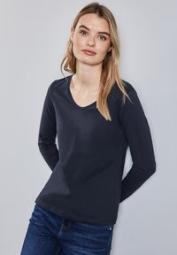 Pitkähihaisen tummansinisen paidan, jossa on pyöreä pääntie ja joka on valmistettu sileästä kankaasta, yhdistetään sinisiin farkkuihin.