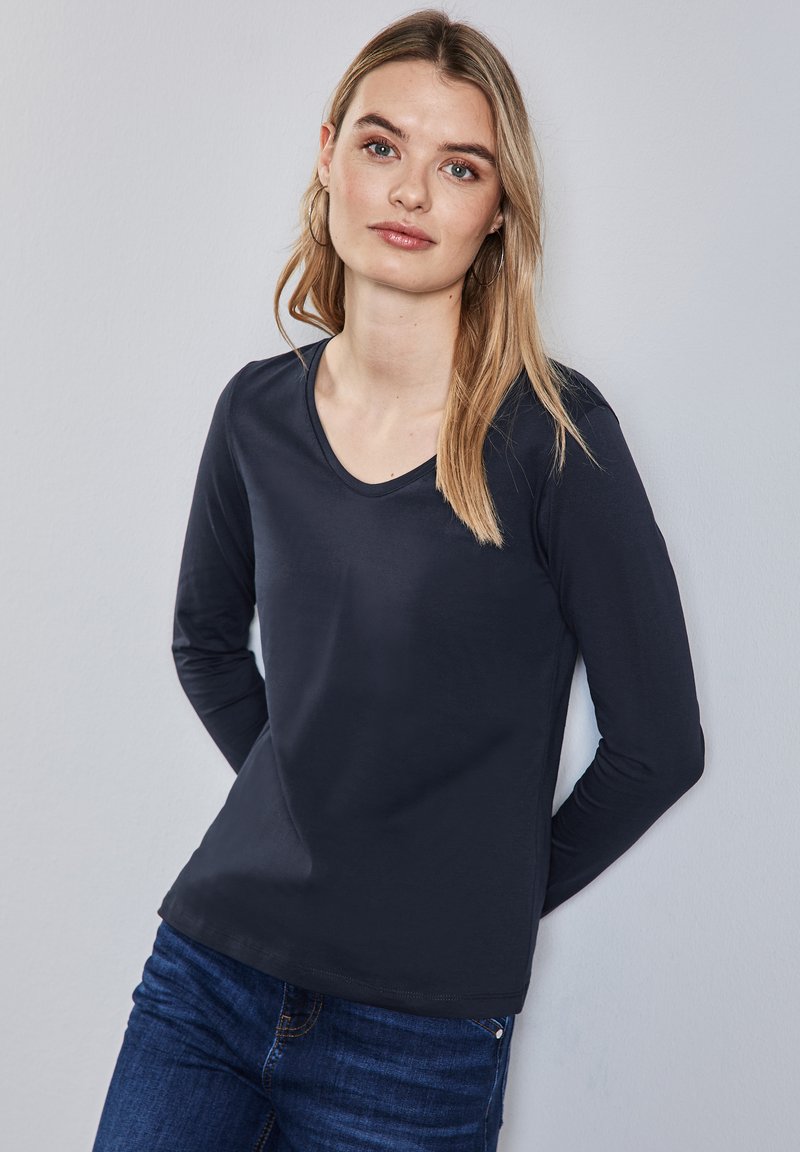 Pitkähihaisen tummansinisen paidan, jossa on pyöreä pääntie ja joka on valmistettu sileästä kankaasta, yhdistetään sinisiin farkkuihin.