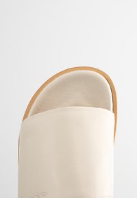 Leather slide sandal with a wide, curved cream strap and a tan sole. The strap features a smooth texture and a minimalistic design.