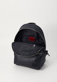 Mochila de cuero negro con un acabado texturizado, cierre de cremallera y etiqueta de logo roja en el interior. Cuenta con un amplio compartimento principal y un bolsillo frontal.