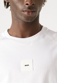 Balta medvilnės t-shirt su kvadratine etikete, kurioje juodu tekstu užrašyta "BOSS", apvaliu kaklu ir sklandžia tekstūra.