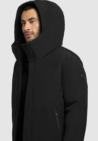 Sort polstret jakke med en stor hætte, udstyret med lynlås og sidelommer. Glat tekstur, minimalistisk design, ingen synlige mønstre.
