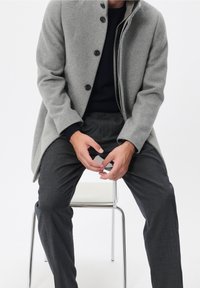 Cappotto di lana grigio chiaro con collo alto e sette bottoni, abbinato a pantaloni grigio scuro. Vestibilità strutturata, con texture liscia e linee pulite.