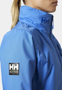 Niebieska wodoodporna kurtka z wysokim kołnierzem, gładką teksturą i logo Helly Tech na rękawie. Zamek błyskawiczny z przodu oraz oddychający materiał.