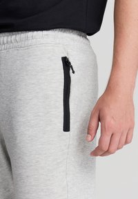 Jasnoszare dresy posiadają teksturowany materiał, elastyczny pas oraz boczną kieszeń z czarnym zamkiem błyskawicznym. Częściowo widoczna ręka.