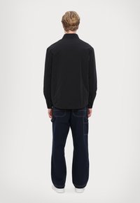 Svart lättviktig jacka med krage, kombinerad med lösa marinblå byxor med flera fickor och kontrasterande sömdetaljer.