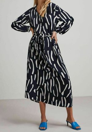 Kobieta w czarnej sukience midi z białymi abstrakcyjnymi paskami i wiązaniem w talii, w połączeniu z jasnoniebieskimi sandałami na obcasie z otwartym palcem.