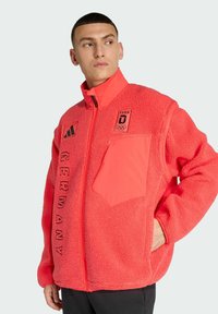 Chaqueta de forro polar roja con cierre de cremallera, que presenta un gran bolsillo en el pecho, texto "GERMANY" y emblema olímpico en el frente.