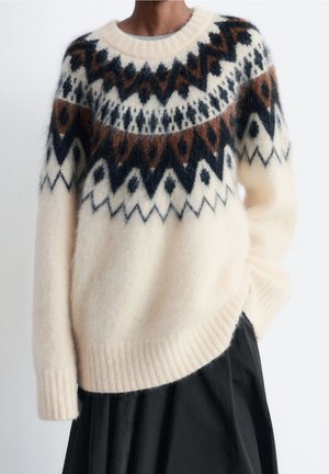 Personne portant un pull en laine crème avec des motifs géométriques noirs et marron sur la poitrine et les épaules, assorti à une jupe noire.