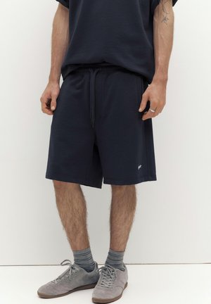 Egy férfi sötétkék megkötős rövidnadrágban, szürke csíkos zokniban és szürke sportcipőben, sima fehér háttér előtt áll.