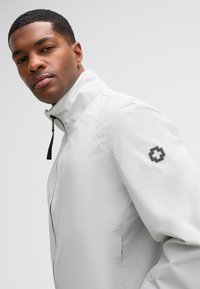 Leichte weiße Jacke mit hohem Kragen, Vorderreißverschluss und schwarzem Logo auf dem linken Ärmel. Glattes Material mit minimalistischen Design.