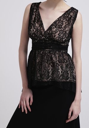 Kvinde iført ærmeløs sort blonder top med V-hals og tætsiddende taljebånd over en ensfarvet sort nederdel, stående mod en ensfarvet baggrund.