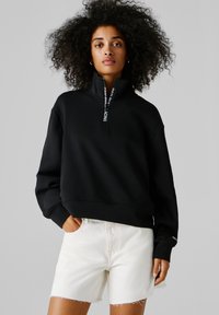 Ženska z kodrastimi lasmi, oblečena v črno majico s četrtinskimi zadrgami in bele kateriške visokopasne kavbojke, na enobarvnem ozadju.