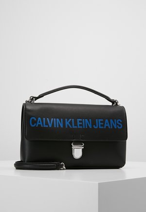 Černá kabelka Calvin Klein Jeans s modrým logem, horním uchycením, stříbrným zapínáním a odnímatelným ramenním popruhem na bílé ploše.