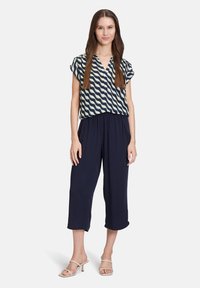 Lekki, granatowy model spodni culotte w połączeniu z krótkim rękawem, wzorzystą bluzką z zielonymi falistymi paskami. Modelka stoi na białym tle.