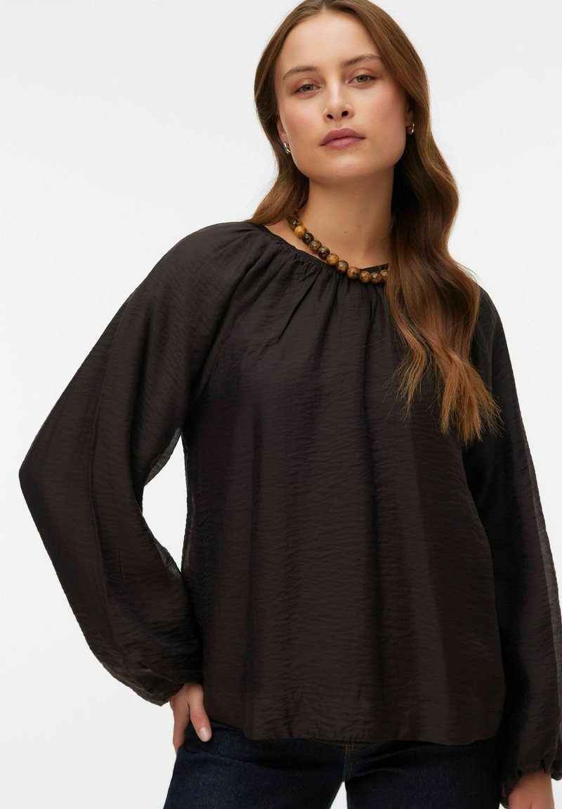 Blusa nera a maniche lunghe con texture e scollatura arricciata. Caratterizzata da una vestibilità comoda e una leggera lucentezza. Abbinata a una collana di perline di legno.