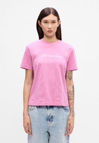 Rozā kokvilnas T-krekls ar apaļu izgriezumu un īsām piedurknēm. Priekšpusē ir balts uzraksts "Calvin Klein Jeans". Brīvā piegriezumā.