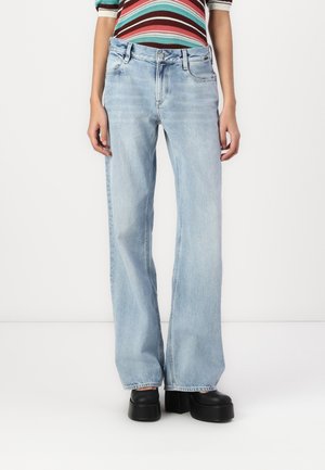 Personne portant un jean à jambes larges bleu clair, des chaussures à plateforme noires et un haut rayé à manches longues, debout devant un fond blanc uni.