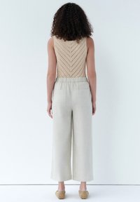 Top in maglia beige con motivo a zigzag, abbinato a pantaloni leggeri a palazzo. La modella indossa scarpe slip-on color nocciola. Sfondo pulito.