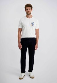 Man staat rechtop in een wit T-shirt met grafisch print, donkere broek en witte sneakers, tegen een effen lichte achtergrond.