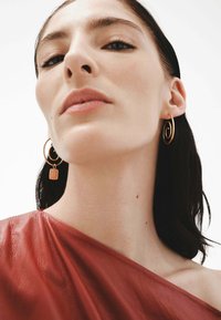 Pendientes de aro dorados con un diseño en espiral y un pequeño charm de candado. El modelo lleva un top de cuero rojo oscuro con una textura suave.