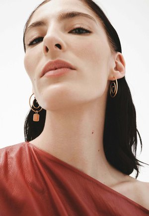 Pendientes de aro dorados con un diseño en espiral y un pequeño charm de candado. El modelo lleva un top de cuero rojo oscuro con una textura suave.