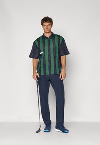 Kortsleeves navy polo med grønne vertikale striper, teksturert mesh-stoff. Sammen med navy bukser og blå treningstid; holder en golfkølle.