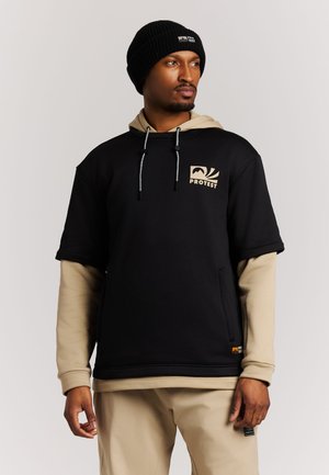 Man draagt een zwarte korte mouw hoodie over een beige lange mouw overhemd en beige broek, met een zwarte gebreide muts op zijn hoofd.