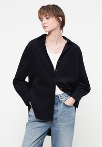 Cămașă din cordă marină cu un guler cu nasturi, buzunar pe piept și mâneci lungi, asociată cu blugi bleu deschis. Detaliu subtil de textură vizibil.