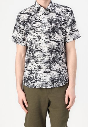 Homme portant une chemise à manches courtes avec des boutons, imprimée de palmiers tropicaux en noir et blanc, et un pantalon vert olive, sur un fond uni.