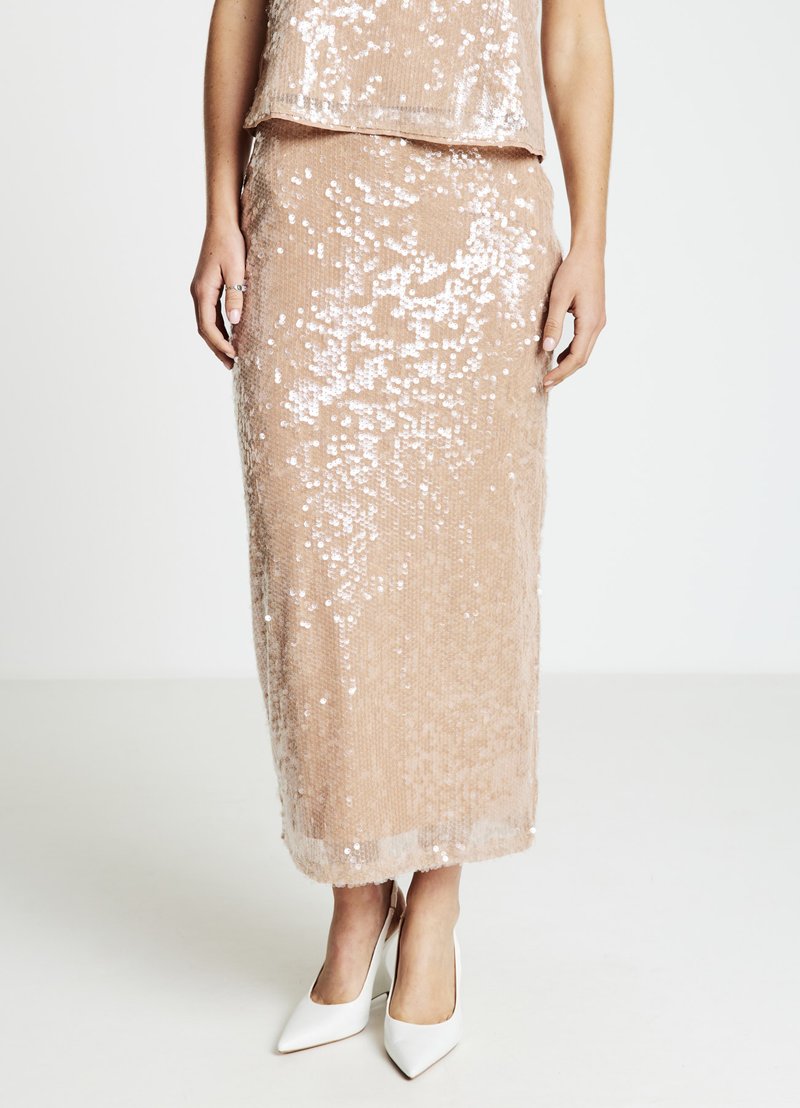 Zweiteiliges Pailletten-Set in sanftem Beige, bestehend aus einem bauchfreien Oberteil und einem langen Rock. Die Pailletten fangen das Licht ein und erzeugen einen schimmernden Effekt. Weiße High Heels.