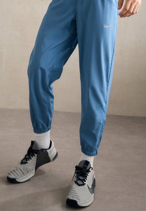 Partie inférieure du corps portant un jogging Nike bleu, des chaussettes blanches et des baskets Nike grises, debout sur un sol clair avec un fond uni.