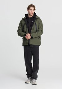 Giacca verde isolata con cappuccio, zip nere e due tasche frontali, abbinata a una felpa nera, jeans neri e sneakers grigie.