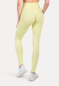 Geel, hooggetailleerde leggings van rekbaar materiaal, met een gladde afwerking en een logodetail op de tailleband. Draagt met witte sneakers.