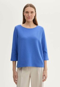 Blaues Baumwollmischungsoberteil mit rundem Ausschnitt und Dreiviertelärmeln, das eine strukturierte Oberfläche und eine lockere, entspannte Passform aufweist.