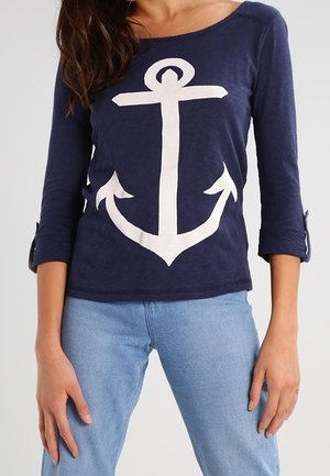 Vrouw draagt een marineblauw shirt met lange mouwen met een grote witte ankerafbeelding en lichtblauwe spijkerbroek.