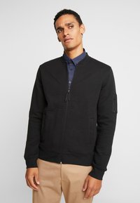 Sort zip-up jakke med ribbet tekstur, båndkrave og sidelommer. Har en lynlåslomme på ærmet. Parret med en mønstret blå skjorte.