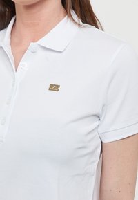 Polo bianco con tessuto testurizzato, colletto con due bottoni e una piccola accentatura dorata sul lato sinistro.