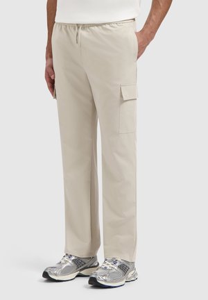 Mann trägt beige Cargo-Hosen mit Seitentaschen und silbernen Laufschuhen, steht mit einer Hand in der Tasche.