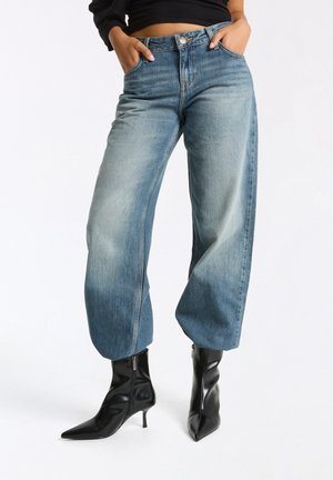 Person trägt hellblaue Jeans mit weitem Bein und schwarze spitze Stiefeletten mit Absätzen und Reißverschlüssen und steht mit den Händen in den Taschen.