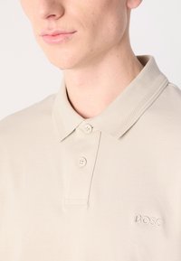Béžová polo košeľa vyrobená z mäkkej bavlny, s klasickým golierom, tromi gombíkmi a jemne vyšitým logom "BOSS" na hrudi.