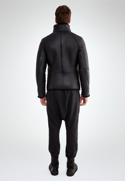 Un uomo visto di spalle che indossa una giacca di pelle nera, pantaloni larghi scuri e scarpe nere alta fino alla caviglia contro uno sfondo neutro.