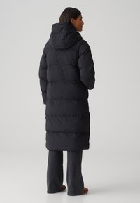 Doudoune noire avec capuche, présentant un design matelassé et une silhouette longue, associée à un jean flare noir et des chaussures rouges.